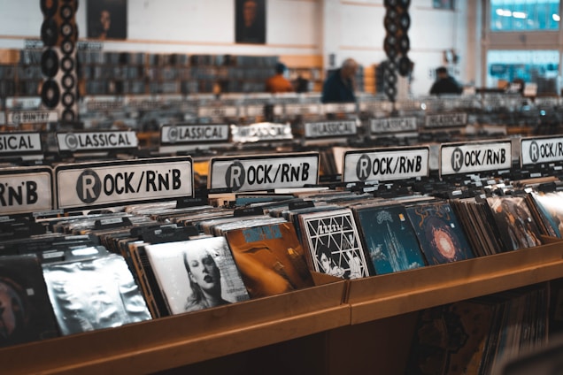 Rows of vinyl records are displayed in a music store, categorized under various genres such as Rock/RnB and Classical. The shelves are filled with album covers featuring diverse artwork. The space is well-lit, suggesting an inviting atmosphere for browsing music enthusiasts.