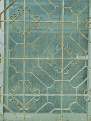 Intricate metal grill work with geometric and floral patterns covers a window, creating an ornate and decorative appearance. The metal is aged, showing signs of wear and rust. The glass behind the grill is slightly dusty, giving a muted view of what lies beyond.