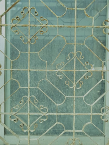 Intricate metal grill work with geometric and floral patterns covers a window, creating an ornate and decorative appearance. The metal is aged, showing signs of wear and rust. The glass behind the grill is slightly dusty, giving a muted view of what lies beyond.