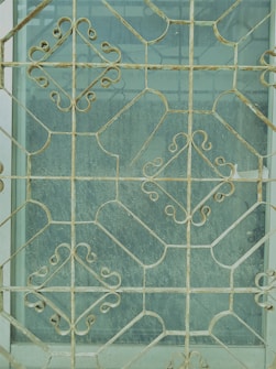 Intricate metal grill work with geometric and floral patterns covers a window, creating an ornate and decorative appearance. The metal is aged, showing signs of wear and rust. The glass behind the grill is slightly dusty, giving a muted view of what lies beyond.