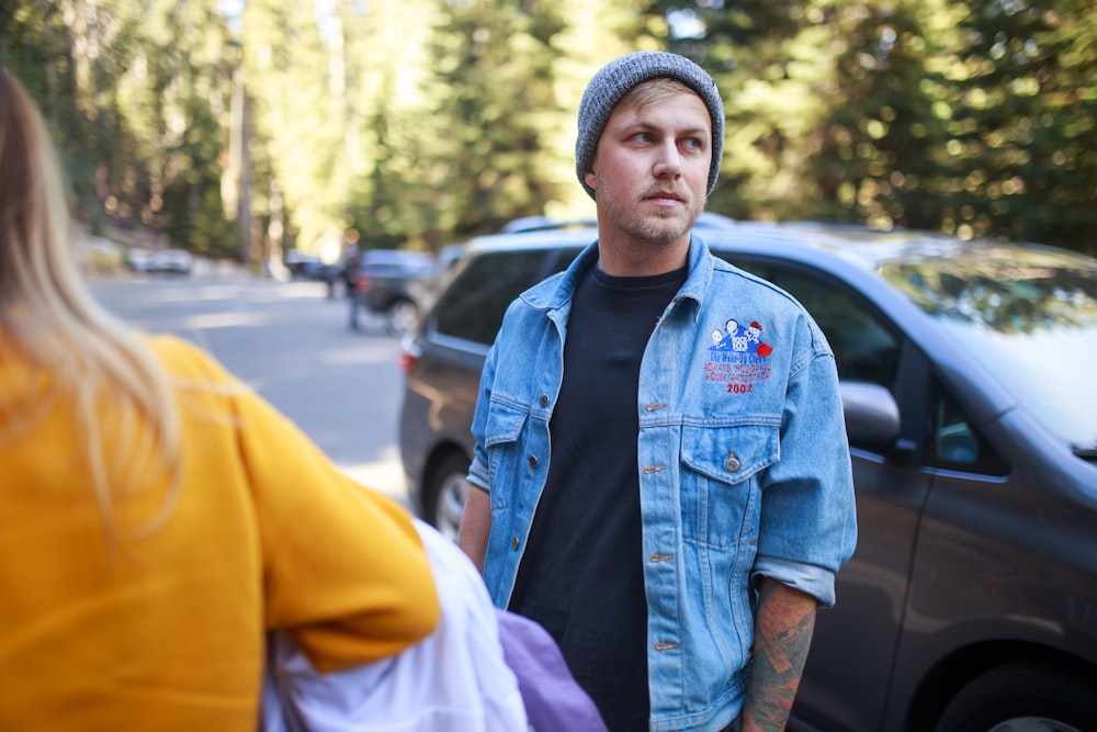 man in blue denim jacket wearing gray knit cap standing near cars during daytime