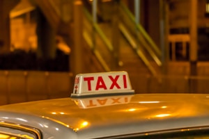 A taxi sign is brightly illuminated against a blurred, warm-toned urban backdrop. The scene suggests nighttime in a city area with lights reflecting off the roof of the vehicle.