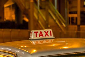 A taxi sign is brightly illuminated against a blurred, warm-toned urban backdrop. The scene suggests nighttime in a city area with lights reflecting off the roof of the vehicle.