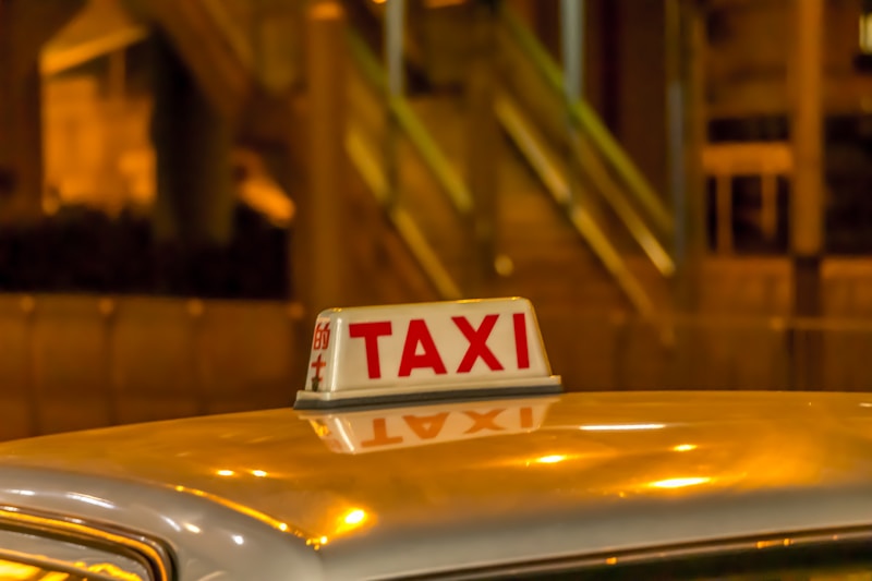 A taxi sign is brightly illuminated against a blurred, warm-toned urban backdrop. The scene suggests nighttime in a city area with lights reflecting off the roof of the vehicle.