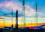 A rocket poised vertically on a launch pad, surrounded by tall metallic structures, likely support towers. In the background, the sky displays a vibrant mixture of colors, including blues, oranges, and yellows, indicating a sunset or sunrise.