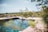 A serene natural pool is surrounded by pink stone slabs, blending into a lush landscape. A crocodile-shaped handrail decorates one side of the pool, while rustic pots are placed on the other. The background features expansive greenery and a clear blue sky, creating a peaceful and tranquil setting.