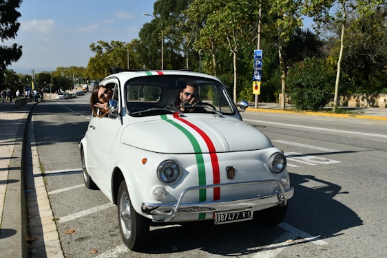 A vintage white car with Italian flag stripes on the hood is parked on the side of a road. A man is seated at the driver’s seat, and a woman leans out of the passenger window, smiling.