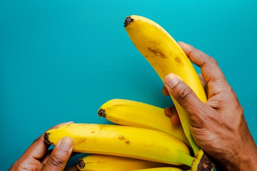 A close-up of hands holding several ripe yellow bananas against a bright blue background. The bananas are slightly spotted, indicating ripeness, and are held gently, showcasing their vibrant color and smooth texture.