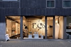 A modern storefront with large glass windows featuring a display of various contemporary lamps and lighting fixtures. The interior is well-lit, showcasing a minimalist and stylish design, with a focus on modern lighting solutions. Visible elements include pendant lamps, floor lamps, and decorative objects arranged neatly. The exterior of the building has a dark-colored facade with vertical paneling.