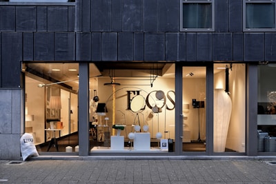 A modern storefront with large glass windows featuring a display of various contemporary lamps and lighting fixtures. The interior is well-lit, showcasing a minimalist and stylish design, with a focus on modern lighting solutions. Visible elements include pendant lamps, floor lamps, and decorative objects arranged neatly. The exterior of the building has a dark-colored facade with vertical paneling.