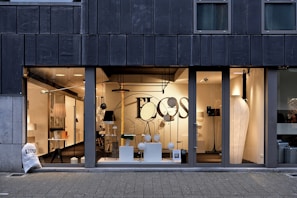 A modern storefront with large glass windows featuring a display of various contemporary lamps and lighting fixtures. The interior is well-lit, showcasing a minimalist and stylish design, with a focus on modern lighting solutions. Visible elements include pendant lamps, floor lamps, and decorative objects arranged neatly. The exterior of the building has a dark-colored facade with vertical paneling.