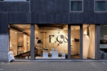 A modern storefront with large glass windows featuring a display of various contemporary lamps and lighting fixtures. The interior is well-lit, showcasing a minimalist and stylish design, with a focus on modern lighting solutions. Visible elements include pendant lamps, floor lamps, and decorative objects arranged neatly. The exterior of the building has a dark-colored facade with vertical paneling.