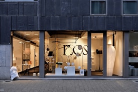 A modern storefront with large glass windows featuring a display of various contemporary lamps and lighting fixtures. The interior is well-lit, showcasing a minimalist and stylish design, with a focus on modern lighting solutions. Visible elements include pendant lamps, floor lamps, and decorative objects arranged neatly. The exterior of the building has a dark-colored facade with vertical paneling.