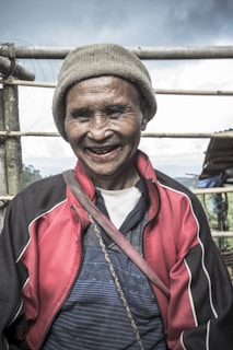 An elderly person is smiling warmly. They are wearing a beige knit hat, a red and black jacket with white accents, and a striped blue shirt. A strap crosses their body, and they stand outdoors with a bamboo fence and cloudy sky in the background.