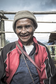 An elderly person is smiling warmly. They are wearing a beige knit hat, a red and black jacket with white accents, and a striped blue shirt. A strap crosses their body, and they stand outdoors with a bamboo fence and cloudy sky in the background.