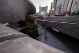 A modern urban landscape with sleek, curved architecture juxtaposed against tall, rectangular skyscrapers. The area is bustling with people on a wide walkway, and there is greenery in the form of a few trees and bushes. Large digital screens and signs are visible on the buildings.