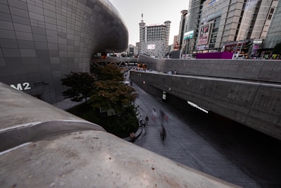 A modern urban landscape with sleek, curved architecture juxtaposed against tall, rectangular skyscrapers. The area is bustling with people on a wide walkway, and there is greenery in the form of a few trees and bushes. Large digital screens and signs are visible on the buildings.