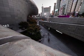 A modern urban landscape with sleek, curved architecture juxtaposed against tall, rectangular skyscrapers. The area is bustling with people on a wide walkway, and there is greenery in the form of a few trees and bushes. Large digital screens and signs are visible on the buildings.