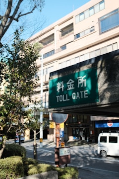 A green sign with white text reading 'Toll Gate' in both English and Japanese hangs in an outdoor urban setting. Behind the sign are buildings with multiple windows, a street, a parked van, and some greenery including trees and bushes.