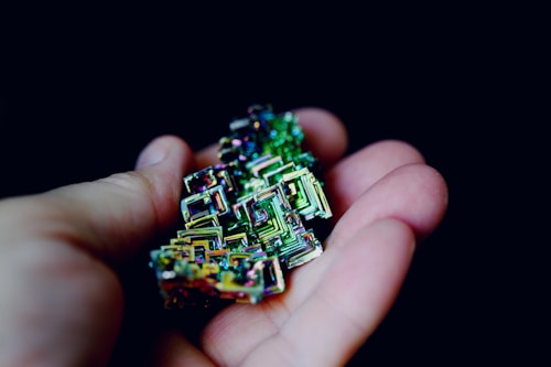A hand gently holds a vibrant bismuth crystal with complex geometric shapes. The crystal displays a range of iridescent colors, adding a mesmerizing quality to the intricate formations.