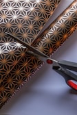 A pair of scissors is cutting through decorative paper with a geometric pattern featuring gold star-like shapes on a black background. The scissors have black handles with red accents, and the paper lies on a flat white surface.