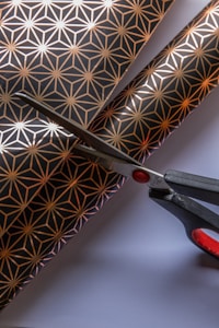 A pair of scissors is cutting through decorative paper with a geometric pattern featuring gold star-like shapes on a black background. The scissors have black handles with red accents, and the paper lies on a flat white surface.
