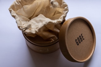A round cardboard box is partially open, revealing a beige, drawstring fabric pouch inside. The box is labeled with the brand name 'Gobe' on the lid, which is placed ajar next to the container. The setting appears to be minimalistic with a neutral background.