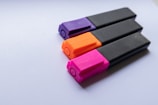 Three highlighters are arranged in a parallel fashion on a white surface. The highlighter caps are colored purple, orange, and pink while their bodies are black.