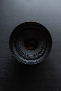 A close-up view of a camera lens centered on a dark textured background. The lens has visible text around its outer edge, indicating its make and specifications.
