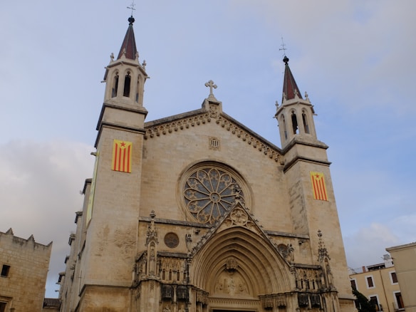 A large, ornate cathedral with two tall spires, each topped with a pointed roof and cross. The facade features intricate stone carvings framing a large circular stained-glass window at the center, above an arched entrance. Flags with red and yellow stripes and stars are displayed on either side of the building.