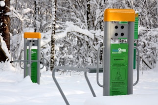 Outdoor gym equipment is installed in a snowy forest, featuring exercise stations with instructional panels. The equipment has gray metal supports with bright yellow tops and green information panels. Snow covers the ground and trees, creating a serene winter setting.