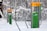 Outdoor gym equipment is installed in a snowy forest, featuring exercise stations with instructional panels. The equipment has gray metal supports with bright yellow tops and green information panels. Snow covers the ground and trees, creating a serene winter setting.