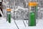 Outdoor gym equipment is installed in a snowy forest, featuring exercise stations with instructional panels. The equipment has gray metal supports with bright yellow tops and green information panels. Snow covers the ground and trees, creating a serene winter setting.