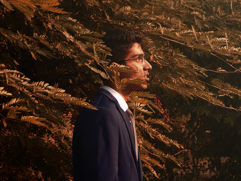 The image depicts a young man standing in front of a bush with brown leaves, wearing a blue suit and tie. The man is looking to his left, suggesting he may have noticed something or someone out of frame. The background features a blurred tree branch, adding depth to the scene and creating an atmosphere of mystery.