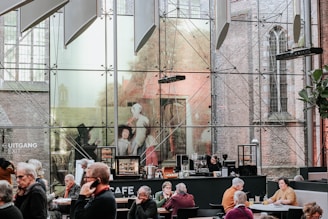 A modern café with large glass walls displaying artwork and allowing a view of a brick building exterior. Several people are seated at tables, engaged in conversation or using their phones, while a woman stands behind the counter, possibly preparing drinks. The ambiance blends contemporary and historical architecture.