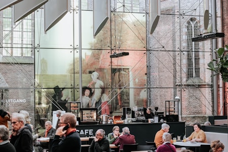 A modern café with large glass walls displaying artwork and allowing a view of a brick building exterior. Several people are seated at tables, engaged in conversation or using their phones, while a woman stands behind the counter, possibly preparing drinks. The ambiance blends contemporary and historical architecture.