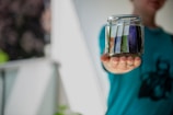 A person is holding a glass jar filled with neatly arranged banknotes. The focus is on the jar and the hand, with the background blurred out. The person is wearing a teal colored shirt with a dark pattern on it.