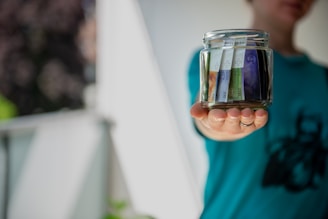 A person is holding a glass jar filled with neatly arranged banknotes. The focus is on the jar and the hand, with the background blurred out. The person is wearing a teal colored shirt with a dark pattern on it.