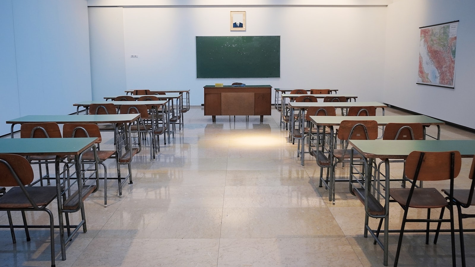 Empty classroom with student desks, chairs, teacher's desk, chalkboard, and portrait on wall