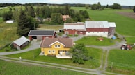 A rural landscape featuring a farmhouse with a yellow facade surrounded by green fields and trees. Several outbuildings with red and gray roofs are visible, along with a red tractor and farming equipment on the grass. A dirt path runs through the property, and electrical poles are present. The background shows rolling hills and additional wooded areas.