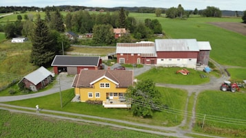 A rural landscape featuring a farmhouse with a yellow facade surrounded by green fields and trees. Several outbuildings with red and gray roofs are visible, along with a red tractor and farming equipment on the grass. A dirt path runs through the property, and electrical poles are present. The background shows rolling hills and additional wooded areas.