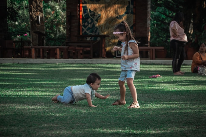 Two young children are playing on a grassy area. One child is crawling on the ground while the other stands nearby, both dressed casually. In the background, adults are sitting and observing the children, surrounded by trees and outdoor furniture.