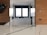 A modern kitchen with sleek white cabinets, wooden wall panels, and a large central window. The room features a minimalist design with glossy surfaces and a marble countertop. A sink is located beneath the window, and there are reflections on the polished surfaces.