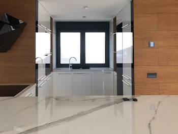 A modern kitchen with sleek white cabinets, wooden wall panels, and a large central window. The room features a minimalist design with glossy surfaces and a marble countertop. A sink is located beneath the window, and there are reflections on the polished surfaces.