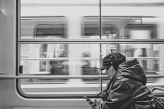 A person wearing a cap and winter jacket is engrossed in their mobile phone while sitting inside a moving train. The blurred motion outside the window suggests the train is moving swiftly past urban scenery.