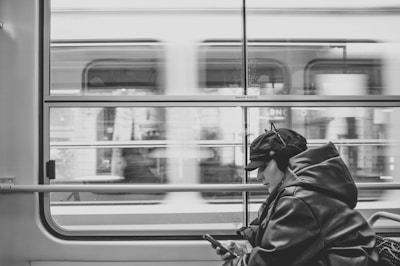 A person wearing a cap and winter jacket is engrossed in their mobile phone while sitting inside a moving train. The blurred motion outside the window suggests the train is moving swiftly past urban scenery.