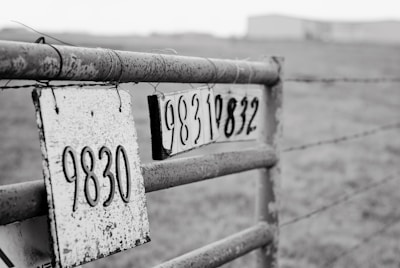 A close-up of a metal gate with two weathered signs displaying numbers 9830 and 9832. The gate is rusty and situated in a grassy area with a blurred background, suggesting an outdoor rural setting.