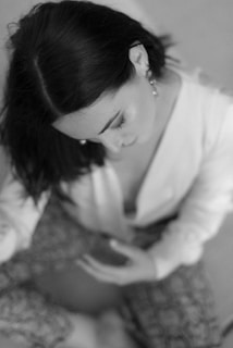 A black and white photograph featuring a person with dark hair looking downwards. They are wearing a light-colored top and patterned pants. The image has a soft focus and a gentle blur, emphasizing the pensive or introspective mood.