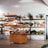 A small shop interior with wooden shelves full of potted plants of various sizes and types. The shelves also hold other items like hats and jars. In front, there's a wooden display cart with more products, including fabric items and small boxes, against a plain wall.