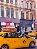 A busy urban street scene featuring a yellow NYC taxi with an advertisement on top. The street is lined with historic buildings housing stores, including Calvin Klein and Ray-Ban, with sale signs displayed in the windows. Multiple pedestrians and cars are visible, contributing to the bustling atmosphere.